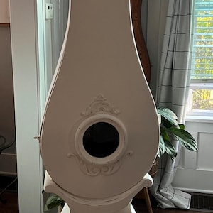 May include: A tall, off-white antique grandfather clock. The clock has a round face with black Roman numerals and ornate hands. The body features a decorative circular element and sits on a pedestal base. The clock is a classic piece of home decor.