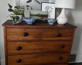 May include: A wooden dresser with five drawers and a white lamp with a white lampshade on top. The dresser has a mirror above it and a plant in a white pot on the left side. There are two blue and white bowls on the dresser.