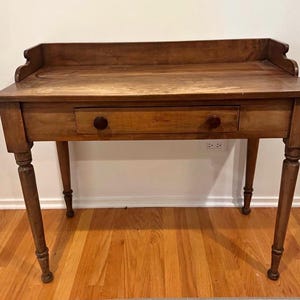 May include: A wooden desk with a warm, medium-brown finish. It features a single drawer with two round knobs and four turned legs. The desk has a raised back edge and is set against a white wall and hardwood floor.