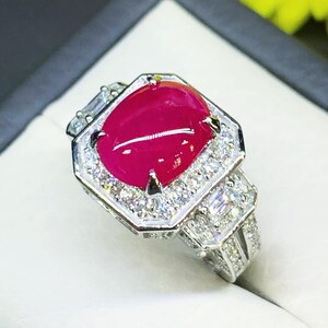 May include: A silver-toned ring featuring a large, oval, cabochon-cut vibrant red gemstone at the center. The stone is surrounded by a halo of small, clear, round-cut stones, with additional baguette-cut stones set along the band's shoulders.