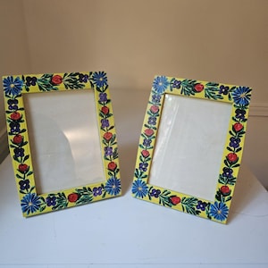 May include: Two yellow picture frames with floral designs. The frames have a rectangular shape with a yellow border. The floral designs include red, blue, and purple flowers and green leaves. The frames are placed on a white surface.