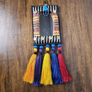 May include: A rectangular wall mirror framed in black and white striped fabric with red and yellow patterned side panels. The mirror is decorated with small gold-colored metal coins and a blue bead at the top, featuring five long, colorful tassels.