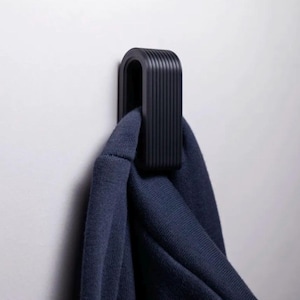 May include: A black, modern wall hook with a ribbed design, holding a navy blue garment. The hook is mounted on a white wall. The design is minimalist and functional.