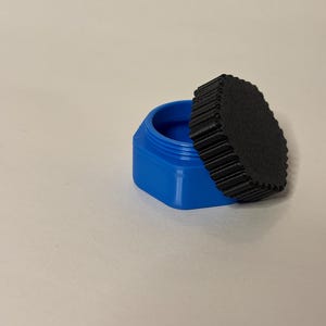 May include: A small, open, blue hexagonal container with a black, circular lid. The lid has a ridged edge and is partially covering the container. The interior of the container is also blue.
