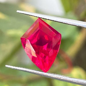 Könnte beinhalten: Ein leuchtend roter, drachenförmiger loser Edelstein mit facettierter Oberfläche, der von einer silberfarbenen Metallpinzette gehalten wird. Der Stein weist scharfe geometrische Kanten und eine brillante, durchscheinende Klarheit auf.