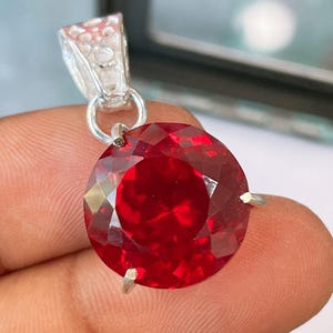 Könnte beinhalten: Ein runder, facettierter, tiefroter Edelsteinanhänger in einer silberfarbenen Metallfassung mit Krappen. Der kreisförmige Stein weist einen Brillantschliff mit mehreren reflektierenden Flächen auf und ist an einer verzierten, strukturierten Metallöse befestigt.