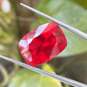 以下が含まれることがあります： シルバーカラーの金属製ピンセットで保持された、鮮やかなクッションカットの赤い宝石。石の表面はファセットカットされており、光を反射して、透明で磨かれた結晶構造全体に深く飽和した深紅の色合いを映し出しています。