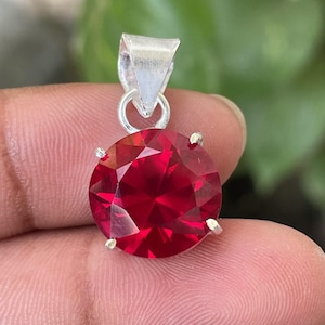 Könnte beinhalten: Ein runder, facettierter, tiefroter Edelsteinanhänger in einer silberfarbenen Metallfassung mit Krappen. Der kreisförmige Stein ist im Brillantschliff mit mehreren reflektierenden Flächen geschliffen und an einer dreieckigen Metallöse befestigt.