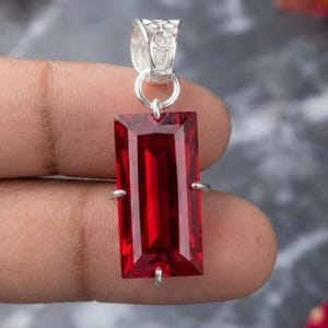 May include: A rectangular, deep red gemstone pendant set in a silver-toned metal frame. The pendant features a faceted, elongated rectangular stone held by four small prongs, attached to a decorative, textured silver-toned bail at the top.