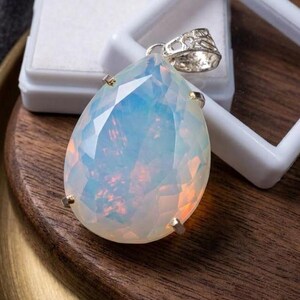 May include: A pear-shaped, faceted opal pendant featuring iridescent blue and orange flashes. The gemstone is secured in a silver-toned metal setting with a decorative, filigree-style bail at the top, showcasing a translucent, milky appearance.