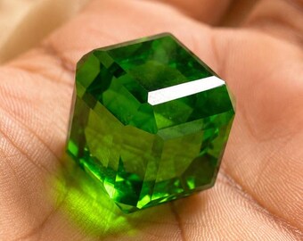 May include: A vibrant, transparent, emerald-green gemstone cut into a precise cube shape. The stone features sharp, clean edges and polished facets that catch the light, creating a brilliant, reflective surface with deep green hues throughout.