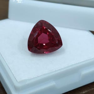 Könnte beinhalten: Ein facettierter, im Triillantschliff geschliffener roter Edelstein mit tiefroter Farbe und hoher Reinheit, der auf der weißen Schaumstoffeinlage einer offenen, quadratischen weißen Kunststoff-Präsentationsbox liegt. Der Stein zeigt viele Facetten.