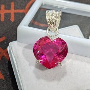 May include: A heart-shaped, faceted hot pink gemstone pendant set in a silver-toned metal prong setting. The pendant features a decorative, filigree-style silver bail at the top, designed to hold a chain, and is presented in a white jewellery box.