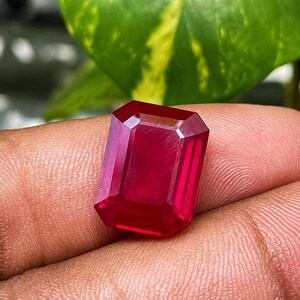 May include: A vibrant, deep red gemstone with a rectangular emerald cut, featuring sharp, clean facets that reflect light across its polished surface. The stone displays a rich, saturated crimson hue throughout its transparent, crystalline structure.