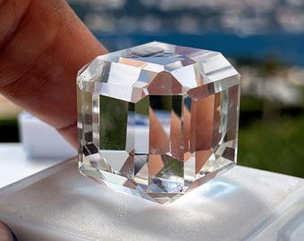 May include: A clear, colorless, faceted gemstone cut into a cube shape. The stone features multiple flat, polished surfaces that reflect light, creating internal refractions. It is displayed resting on a white foam insert within a small box.