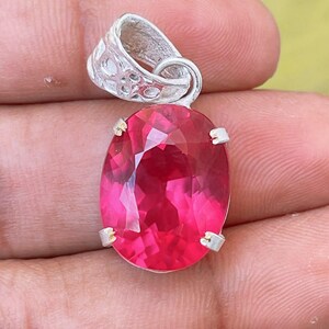 Könnte beinhalten: Ein Anhänger mit einem facettierten, ovalen, leuchtend pinkfarbenen Edelstein in einer silberfarbenen Metallfassung mit Krappen. Der obere Teil des Anhängers besteht aus einer dekorativen, strukturierten silberfarbenen Öse mit filigranem Muster.