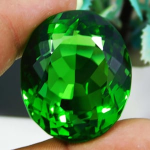 Könnte beinhalten: Ein großer, facettierter, oval geformter Edelstein in leuchtendem Smaragdgrün. Der Edelstein wird zwischen zwei Fingern gehalten, wodurch sein Brillantschliff und seine reflektierenden Oberflächen zur Geltung kommen. Die Facetten erzeugen ein schillerndes Licht- und Schattenspiel.