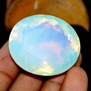May include: A large, oval-shaped gemstone with a faceted surface. The gem displays a soft, iridescent play of colors, primarily in shades of blue, white, and yellow. The stone is held in a hand against a dark background.