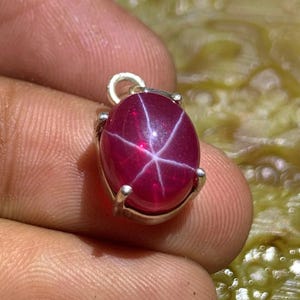 Könnte beinhalten: Ein ovaler, polierter Rubin-Anhänger mit Sterneneffekt. Der tiefrote Edelstein ist in eine silberfarbene Metalleinfassung gefasst und hat eine kleine Schlaufe für eine Kette. Das Sternmuster ist weiß und auf der Oberfläche sichtbar.