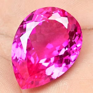 Könnte beinhalten: Ein facettierter, birnenförmiger Edelstein in einem leuchtenden Pinkton. Die Oberfläche des Edelsteins reflektiert das Licht und zeigt seine komplizierten Schnitte und Winkel. Der Stein hat eine tiefe, gesättigte rosa Farbe mit einem polierten, glänzenden Aussehen.