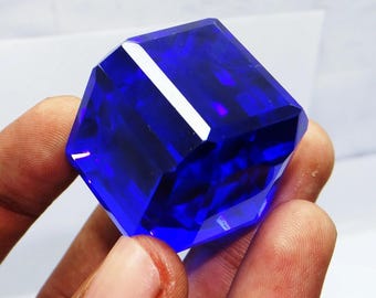 May include: A faceted, deep blue gemstone in a cube shape. The gem is held in a hand, showcasing its brilliant cut and reflective surfaces. The gem's facets catch the light, creating a dazzling display.
