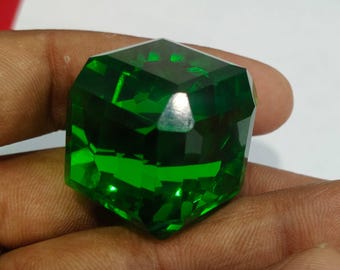 May include: A faceted, emerald-green gemstone with a geometric shape. The gem is held in a hand, showcasing its brilliant cut and reflective surfaces. The gem's deep green color is highlighted by the light.