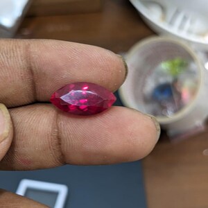 Könnte beinhalten: Ein facettierter Edelstein im Marquiseschliff mit tiefroter Farbe. Der Stein wird zwischen Fingern gehalten, wodurch seine längliche Form und die brillanten Facetten hervorgehoben werden. Der Hintergrund ist unscharf, mit Andeutungen anderer Objekte.