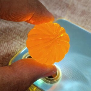 Könnte beinhalten: Ein durchscheinendes, orangefarbenes, blütenförmiges Objekt mit detaillierten, blütenblattartigen Rippen. Das Objekt wird zwischen zwei Fingern gehalten, mit einem verschwommenen Hintergrund eines Telefons und einer gemusterten Oberfläche.