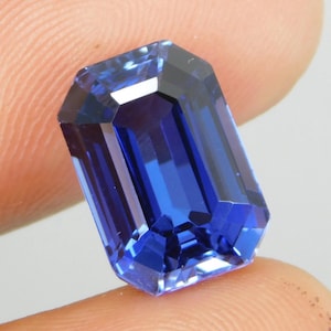 Puede incluir: Una piedra preciosa de zafiro tallada en forma rectangular. La gema es de un azul profundo y vibrante con una superficie pulida, que refleja la luz. El corte presenta una serie de facetas paralelas, creando una brillante exhibición de color y luz. La piedra preciosa se sostiene entre dos dedos.