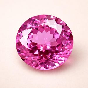 Könnte beinhalten: Ein runder, facettierter rosa Edelstein mit Brillantschliff. Der Edelstein zeigt einen lebendigen, satten rosa Farbton und ist vor einem hellen Hintergrund platziert. Die Facetten erzeugen ein Spiel von Licht und Schatten, das seinen Glanz verstärkt.