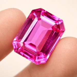 Könnte beinhalten: Ein großer, rechteckig geschliffener rosa Edelstein mit vielen Facetten. Der Edelstein hat eine lebendige, gesättigte Farbe und einen hohen Reinheitsgrad, wobei mehrere Facetten das Licht reflektieren. Der Edelstein wird zwischen zwei Fingern gehalten.
