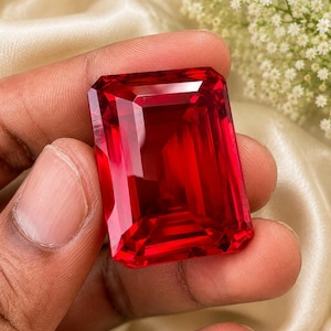 Könnte beinhalten: Ein großer, rechteckiger Edelstein im Smaragdschliff mit einer tiefen, leuchtend roten Farbe. Der Stein weist präzise, facettierte Kanten auf, die eine klare, reflektierende Oberfläche erzeugen und seine hohe Reinheit sowie den intensiven Farbton unterstreichen.