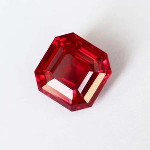 Könnte beinhalten: Ein facettierter, achteckiger, tiefroter Edelstein. Der Edelstein hat einen Brillantschliff mit mehreren Facetten, die das Licht reflektieren. Der Edelstein ist vor einem weißen Hintergrund platziert, der seine leuchtende Farbe und Klarheit hervorhebt. Der Edelstein scheint ein Rubin zu sein.