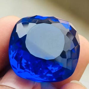 Puede incluir: Una gema facetada, de talla cojín, en un vibrante tono azul profundo. La superficie de la gema refleja la luz, mostrando sus intrincados cortes y facetas pulidas.