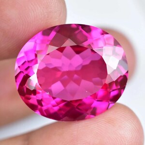 Könnte beinhalten: Ein großer, facettierter, oval geformter Edelstein in einem leuchtenden Pinkton. Der Stein wird zwischen zwei Fingern gehalten, wodurch sein Brillantschliff und seine reflektierenden Oberflächen zur Geltung kommen. Die Facetten des Steins erzeugen ein schillerndes Spiel von Licht und Farbe.