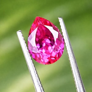 Könnte beinhalten: Ein facettierter, birnenförmiger Edelstein in Rosa- und Magentatönen, gehalten von einer silbernen Pinzette. Der Schliff des Edelsteins zeigt mehrere Facetten, die das Licht reflektieren. Der Hintergrund ist ein weiches, verschwommenes Grün.