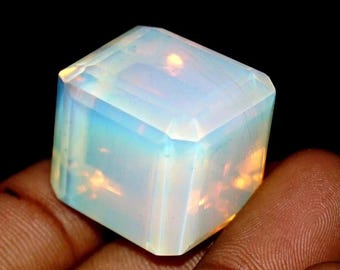 May include: A faceted, cube-shaped opal gemstone. The gem displays a milky white base color with iridescent flashes of blue and gold. The stone is held in a hand against a black background.