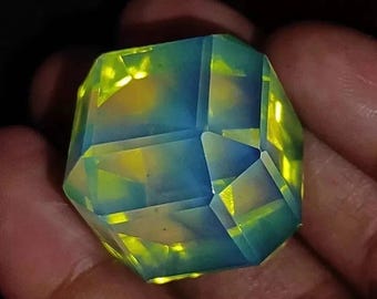 May include: A faceted, iridescent gemstone with a complex geometric design. The gem features a blend of blue, yellow, and green hues, creating a luminous effect. The crystal-like structure is held in a hand, showcasing its intricate cuts and polished surfaces.