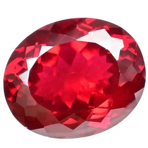 Puede incluir: Una gema roja oscura facetada, de forma ovalada. La gema tiene un corte brillante con numerosas superficies reflectantes, creando un efecto brillante. La piedra se encuentra sobre un fondo blanco liso, resaltando su color y claridad.