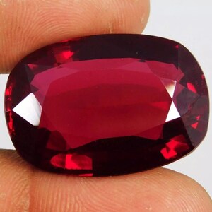 Könnte beinhalten: Ein facettierter, oval geformter Edelstein in tiefroter Farbe. Die Oberfläche des Edelsteins reflektiert das Licht und zeigt seinen Schliff und seine Klarheit. Der Stein wird zwischen Fingern gehalten, was seine Größe und Farbe hervorhebt. Dieser Edelstein ist für Schmuck.