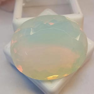 Könnte beinhalten: Ein großer, facettierter, oval geformter Edelstein mit milchig-opaleszierendem Aussehen. Der Edelstein hat eine weiche, perlmuttartige weiße Farbe mit subtilen rosa und gelben Farbtönen, vor einem weißen Hintergrund.