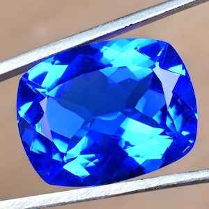 Puede incluir: Una gema vibrante, de talla cojín, en un tono azul profundo y saturado. Las facetas reflejan la luz, creando un brillo brillante. Sujeta por pinzas plateadas, la forma de la gema es un cuadrado redondeado, ideal para joyería.