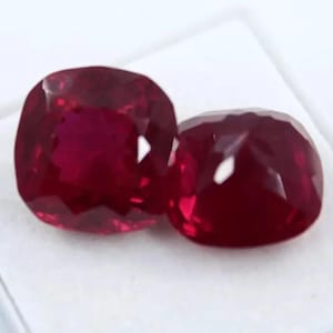 Könnte beinhalten: Zwei kissenförmig geschliffene Rubine mit tiefroter Farbe. Die Edelsteine sind facettiert und reflektieren das Licht, wodurch ihre Klarheit zur Geltung kommt. Die Rubine sind vor einem weißen Hintergrund platziert, was ihre Farbe und Brillanz hervorhebt. Ideal für die Schmuckherstellung.