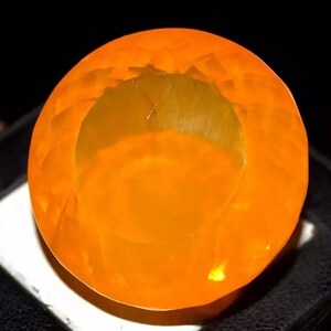 Puede incluir: Una gema redonda y facetada de color naranja con un centro hueco. La gema es de un color naranja vibrante y translúcido, con una superficie pulida que refleja la luz. La gema está sobre un fondo oscuro.