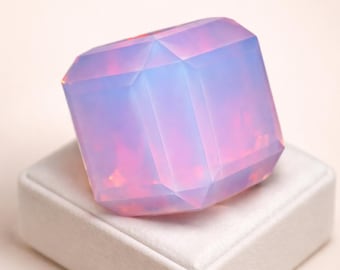 May include: A faceted, rectangular gemstone with a soft, opalescent glow. The gem displays a gradient of colors, transitioning from a pale blue to a vibrant pink. It rests on a white, square display.