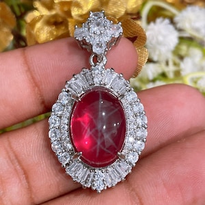 May include: A silver-toned pendant featuring a large, oval-shaped, deep red cabochon gemstone with a visible star pattern. The center stone is surrounded by a double halo of clear, sparkling baguette and round-cut white crystals.