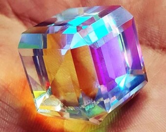 May include: A faceted, clear crystal cube with iridescent color reflections of blue, purple, and yellow. The cube is held in a hand, showcasing its geometric shape and light refraction. This decorative crystal is ideal for display.