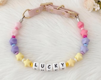 Puede incluir: Un collar para perros en colores pastel con la palabra "LUCKY" escrita con cuentas de letras blancas. El collar presenta una correa de cuero rosa con herrajes dorados y está adornado con cuentas en forma de estrella en varios colores.