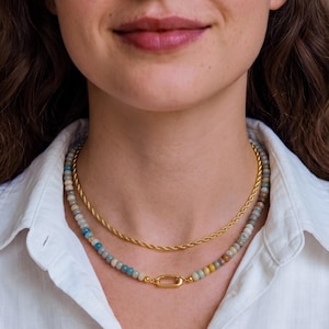 Op de afbeelding: Een gelaagde kettingenset bestaande uit een goudkleurige gedraaide ketting en een tweede streng met kleine, veelkleurige kralen in blauwe, grijze en beige tinten. De kralenketting heeft een centrale goudkleurige, ovale karabijnsluiting.