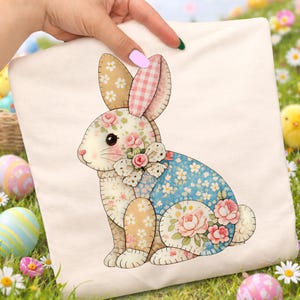 Puede incluir: Un cojín crema con temática de Pascua con un diseño de conejito de patchwork. El conejito tiene orejas, cuerpo y pies con estampado floral y cuadros. Está adornado con rosas rosas y un lazo. El cojín está sostenido por una mano.